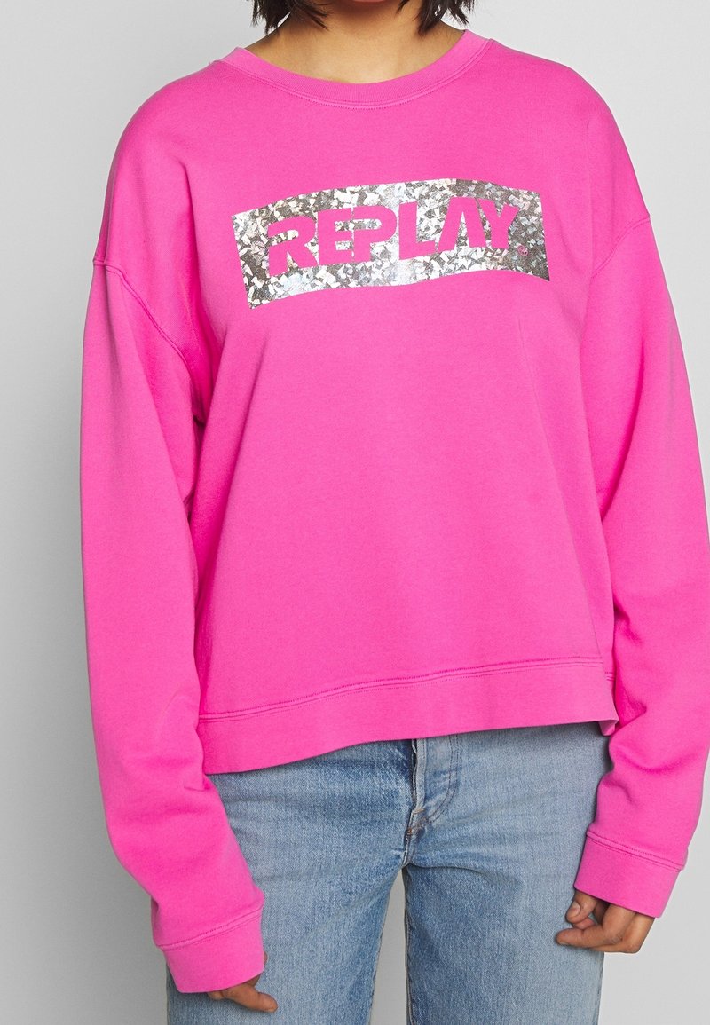 Sudadera de color rosa brillante con un corte recortado, que presenta un logo plateado en lentejuelas "REPLAY" en la parte delantera. Tejido suave y puños acanalados.