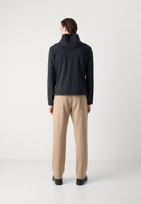 Black lightweight jacket with a hood, featuring smooth texture and fitted design, paired with cream-colored trousers and black shoes.