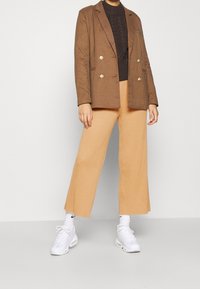 Brun rutigt dubbelknäppt blazer över en mörk tröja, kombinerat med lösa, ribbade beige byxor och vita sneakers.