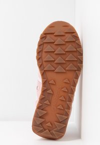 Semelle d'une chaussure rose avec une bande de roulement en caoutchouc marron comportant de larges crampons triangulaires et le nom de la marque « Saucony » embossé au centre.