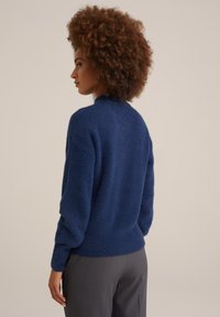 Mörkblå luddig tröja med hög krage, ribbade ärmslut och nederkant. Mjuk textur med en lös passform, kombinerad med grå byxor.