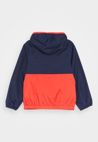 Kapuzensjacke mit der oberen Hälfte in Marineblau und der unteren Hälfte in leuchtendem Rot. Hergestellt aus glattem, leichtem Material mit elastischen Bündchen und Saum.