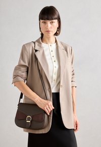 Sac à main marron et bordeaux avec un rabat incurvé et des éléments en or, associé à un blazer beige sur un haut blanc à boutons et une jupe noire.