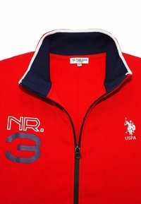Röd zip-up sweatshirt med marinblå krage, vita stripdetaljer och broderade logotyper. Har "NR. 3" i blått på vänster sida. Tillverkad av bomull.