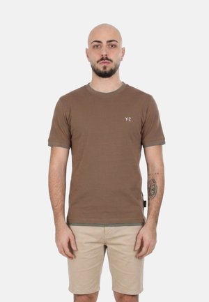 Uomo calvo con barba in piedi di fronte, indossa una maglietta marrone con logo Y-Z e pantaloncini beige, espressione neutra, sfondo bianco uniforme.