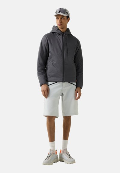 Jonge man met een donker vest met capuchon, lichtgrijze korte broek met ritszakken, witte sokken, sneakers, een honkbalpet en een zonnebril op de pet.