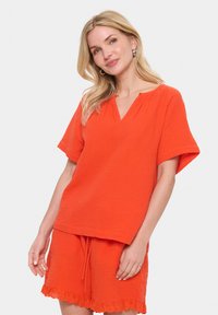 Žiarivý oranžový dvojdielny outfit, ktorý obsahuje top s krátkym rukávom a výstrihom do V a šortky na gumu s detailom čipky. Mäkká, štruktúrovaná tkanina.