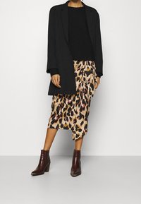 Long blazer noir sur un pull noir, associé à une jupe midi beige à imprimé léopard. Bottines marron à talons avec bouts pointus.