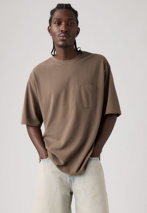 Camiseta de algodón marrón con mangas cortas y un bolsillo en el pecho, combinada con pantalones vaqueros de mezclilla azul claro. Corte sencillo y relajado con textura suave.