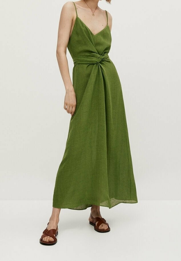 Femme portant une robe maxi verte sans manches avec de fines bretelles et un détail de nœud à la taille, associée à des sandales plates marron.