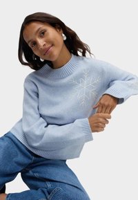 Jasnoniebieski sweter dziany z wysokim kołnierzem i białym haftem w śnieżynki na przodzie i rękawach. W zestawie z niebieskimi dżinsami.