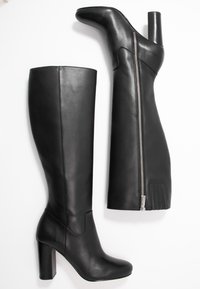 Pár černých kožených kozaček po kolena s blokovými podpatky a viditelným stříbrným zipem na jedné kozačce ležících na bílém pozadí.