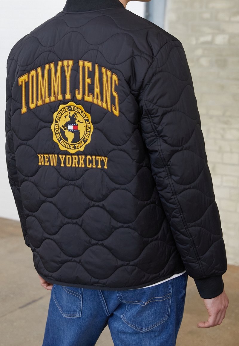 Blouson bomber matelassé noir avec col côtelé, arborant un texte brodé jaune : "TOMMY JEANS" et "NEW YORK CITY", ainsi qu'un accent logo rond.