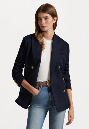 Blazer doppiopetto blu navy con bottoni dorati, taglio sartoriale e colletto a risvolto. Indossato sopra una maglietta bianca e jeans azzurri chiari.