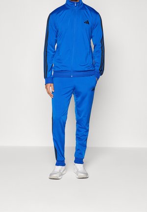 Uomo che indossa una tuta Adidas blu con strisce nere sulle maniche e sui pantaloni, abbinata a sneaker bianche, in piedi davanti a uno sfondo bianco.