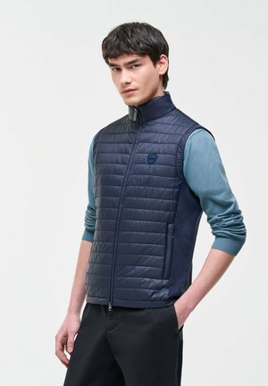 Mladý muž nosí námornícku prešívanú vestu cez modrozelenú košeľu s dlhým rukávom, s jednou rukou v čiernej vrecku nohavíc, na jednofarebnom svetlom pozadí.