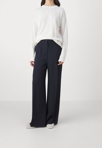 Maglione a maglia crema con collo rotondo e spalle ribassate, abbinato a pantaloni bleu navy a vita alta e gamba ampia, e scarpe di colore chiaro.