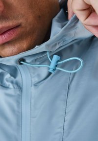 Ljusblå jacka med en slät yta, med en dragsko och ett knäpp som drar åt vid huvan, delvis visad mot hud.