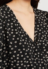 Czarna bluzka kopertowa w kwiatowy wzór z głębokim dekoltem w serek; ozdobiona beżowymi kwiatami i małymi białymi kropkami, wykonana z teksturowanego materiału.
