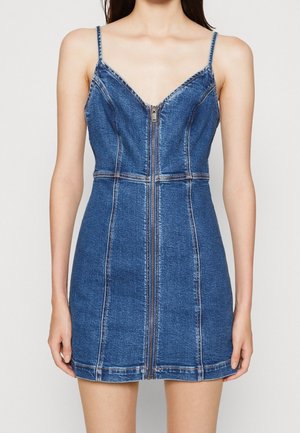Kvinde iført en ærmeløs blå denim mini-kjole med lynlås foran og tynde skulderstropper.