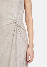 Lśniąca jasnobeżowa sukienka z marszczeniem w talii zdobiona błyszczącymi klejnotami. Tkanina ma fakturowane, faliste wykończenie.