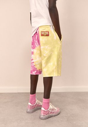 Shorts tie-dye multicolores en jaune et rose avec une écusson portant "GCDS WEAR." Portés avec des chaussettes roses et des baskets translucides.