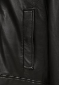 Gros plan sur une veste en cuir noir mettant en avant une poche plate, une texture lisse et des coutures précises le long des bords, soulignant l'artisanat de qualité.