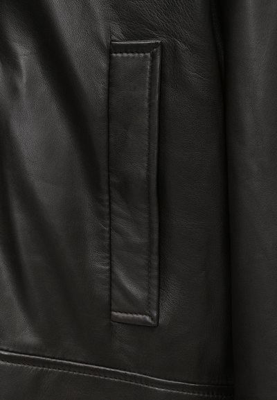 Melnas ādas jakas tuvplāns, kas izceļ plakano kabatu, gludo tekstūru un precīzu šuvju malu, uzsverot kvalitātes meistarību.