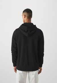 Svart hettegenser med mykt stoff, lange ermer og ribbestrikkede mansjetter. Har en liten logodetalj på ryggen.
