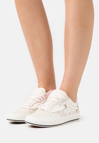 Světlé krémové tenisky s texturovaným materiálem a růžovými akcenty, se šněrovacím designem a perforovanými detaily na špičce.