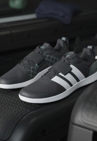 Gray athletic shoes with white stripes, featuring a textured upper and sturdy sole. Includes Velcro strap and geometric designs in teal.