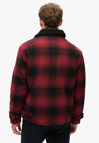 Rød og sort ternet jakke med sherpa-krave, knappede manchetter og en klassisk, struktureret pasform. Mønstrene er bold og jævnt fordelt.