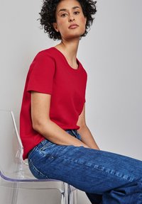 Rode katoenen t-shirt met een ronde hals en korte mouwen, gecombineerd met blauwe spijkerbroek. Model zit op een heldere acrylstoel tegen een effen achtergrond.