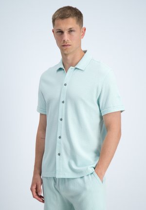 Junger Mann trägt hellblaues, strukturiertes, kurzärmeliges Hemd mit Knöpfen und passende Shorts, steht mit einer Hand in der Tasche vor einfachem Hintergrund.