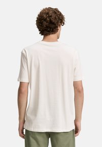 A person with curly hair wears a plain white t-shirt and green pants, viewed from behind against a light gray background.