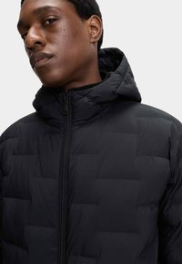 Giacca nera imbottita con chiusura a zip e cappuccio. Presenta un motivo a rombi e una texture liscia. Nessun branding o dettagli visibili.