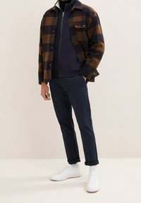 Giacca in flanella a quadri marrone e blu navy, indossata sopra una giacca blu navy con zip, abbinata a pantaloni blu navy e sneakers bianche.
