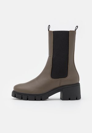 Botte mi-mollet en cuir taupe avec panneau latéral élastique noir, semelle épaisse à crampons, talon bloc et languettes de tirage sur les deux bords supérieurs.