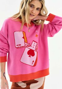 Frau mit blondem Haar, die einen pinken Pullover mit roten Bündchen trägt, auf dem ein Feuerzeug mit Ass-Pik-Karten abgebildet ist, sowie einen braunen und beigen Rock.