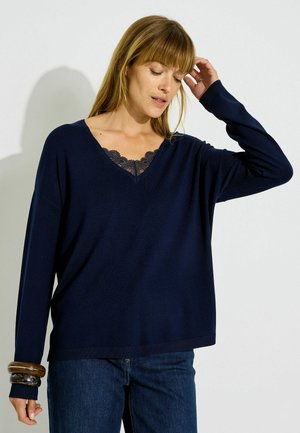 Femme aux cheveux châtain clair portant un pull bleu marine à col en V sur un top en dentelle noire, un jean bleu et des bracelets épais à son poignet gauche.