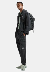 Czarny sportowy zestaw ubrań składający się z kurtki na zamek i spodni typu cargo, zestawiony z jasnoszarymi sneakerami i czarnym plecakiem.