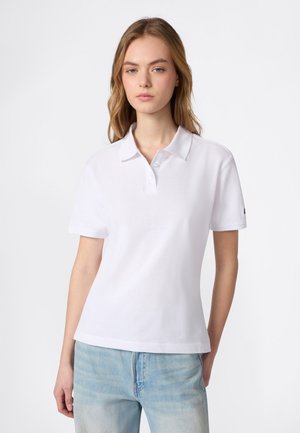 Jeune femme aux cheveux clairs portant un polo blanc uni à manches courtes et un jean bleu clair, debout devant un fond blanc uni.