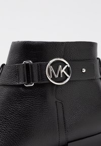 Schwarze Leder-Ankle-Boots mit genarbter Textur, ausgestattet mit einem silbernen, runden MK-Logo und einem verstellbaren Riemen. Glatte, angewinkelte Silhouette.