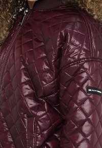 Bordeaux gewatteerde pufferjack met een glanzende afwerking, geribbelde kraag en een logo-patch aan de zijkant met zwart-witte accenten.