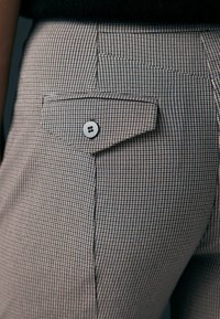 Pantalons à motif pied-de-poule brun et bleu avec une poche arrière dotée d'un détail en bouton argenté. Le matériau semble lisse et bien ajusté.