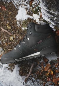 Scarponcino da trekking nero con occhielli in metallo che riposa su terreno innevato e roccioso con foglie secche e rametti.