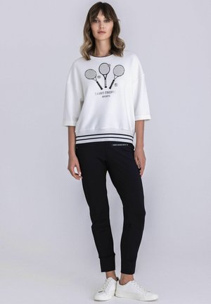 Frau mit weißem Sweatshirt, das eine Grafik mit drei Tennisschlägern und Bällen zeigt, schwarzen Hosen und weißen Turnschuhen, stehend vor einem einfarbigen Hintergrund.