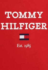 Der rote Baumwollstoff zeigt großen weißen Text "TOMMY HILFIGER", ein kleines mehrfarbig gesticktes Fähnchen und "Est. 1985" in Weiß.