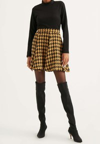 Mujer vistiendo una blusa negra de mangas largas, una falda de pata de gallo amarilla y negra, medias negras transparentes y botas negras de tacón alto hasta la rodilla.