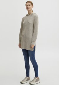 Beige gestrickter Kapuzenpullover mit Rippstruktur, Kordelzugverschluss und seitlichen Schlitzen, kombiniert mit blauen Skinny Jeans und hellen Sneakers.